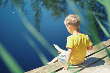 Happy child boy sitting on wooden pier near lake on summy summer day. Toddler kid in yellow t-shirt...