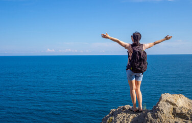 brunette woman in t-shirt with backpack stands back on rocky seaside arms outstretched, and looks out at the sea. Solo summer outdoor activities in fresh air. Concept of Hiking trekking. Copy space