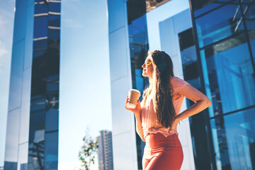 Young sporty fit woman wearing peach sportswear standing near a glass building with paper coffee cup. Bottom view.