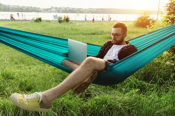 Young successful smart bearded entrepreneur typing on laptop while lying in hammock in park at sunset