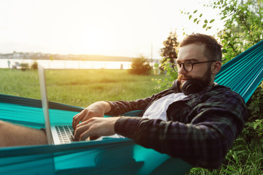 Thoughtful bearded male hipster freelancer developer lying in hammock and working on laptop in park