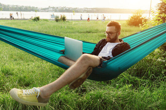 Young successful smart bearded entrepreneur typing on laptop while lying in hammock in park at sunset