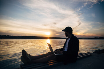 Silhouette of freelancer hipster male in baseball cap working on laptop on riverside in park at sunset.