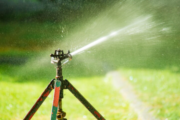 Lawn water sprinkler spraying water over lawn green fresh grass in garden on hot summer day. Automatic watering equipment, lawn maintenance, gardening and Irrigation system. Blurred background. 