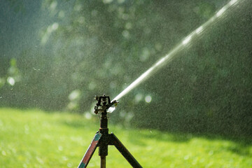 Lawn water sprinkler spraying water over lawn green fresh grass in garden on hot summer day. Automatic watering equipment, lawn maintenance, gardening and Irrigation system. Blurred background. 
