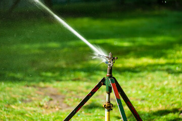 Lawn water sprinkler spraying water over lawn green fresh grass in garden on hot summer day. Automatic watering equipment, lawn maintenance, gardening and Irrigation system. Blurred background. 