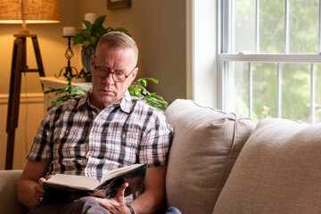 Mature man sits alone, reading a book on living room couch on a rainy day.