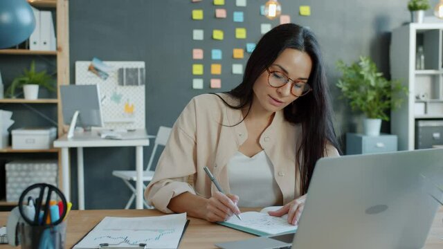 Young businesswoman in elegant clothing and glasses is writing in notebook and using computer smiling in office. Technology and occupation concept.