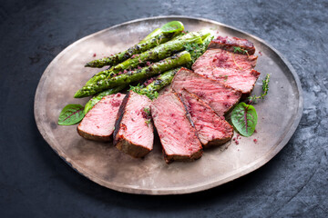 Barbecue dry aged wagyu roast beef steak with green asparagus and lettuce offered as close-up on a rustic modern design plate