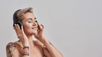 Fototapeta premium Clear Sound. Attractive tattooed woman with pierced nose and short hair in headphones enjoying listening to music isolated over grey background
