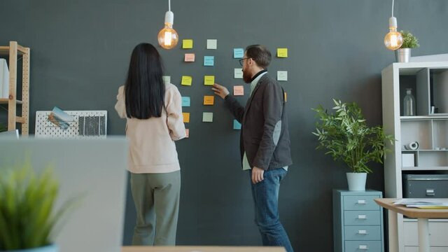 Man and woman are working with sticky notes on wall in office talking sharing business ideas. Communication, creative people and project concept.