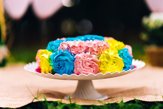 bolo confeitado azul amarelo rosa em boleira branca texturizada com bordas em cima de toalha r&uacute;stica e gramado verde fundo desfocado com bal&otilde;es rosados  