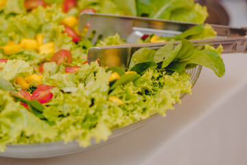 salada de folhas de alface com tomate e milho em refratário de vidro em cima de uma mesa branca e um pegador de inox apoiado 