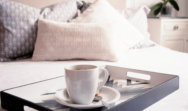 Plano levemente elevado de una taza de cer&aacute;mica blanca con caf&eacute; espumoso sobre una bandeja con estilo y de dise&ntilde;o en una habitaci&oacute;n bien iluminada y sobre una cama de s&aacute;banas blancas