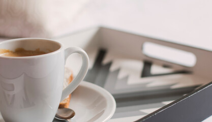 Primer plano frontal de una taza de cerámica blanca con café espumoso sobre una bandeja con estilo y de diseño en una habitación bien iluminada y sobre una cama de sábanas blancas