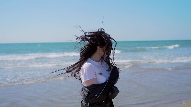 Lifestyle. Portrait of a young attractive sexy and stylish girl in a black leather jacket sunglasses with black hair fluttering in the wind. happy rejoices smiling and spinning around. background sea
