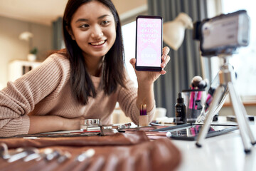 Fototapeta premium Download it. Beauty blogger woman filming, advertising app on camera, holding smartphone. Makeup influencer asian girl recording cosmetics product review at home