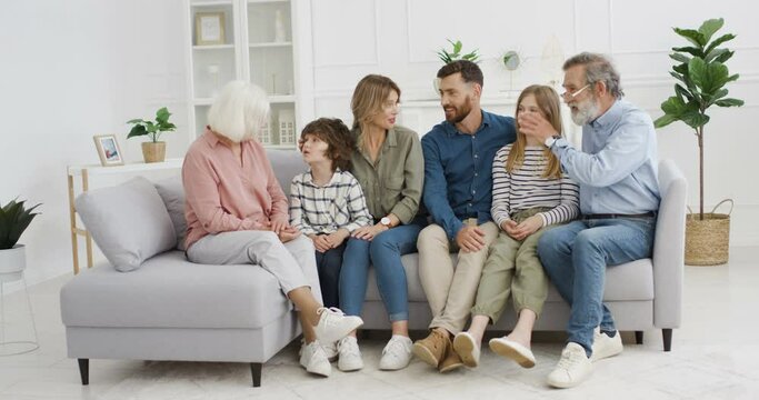 Caucasian happy family with kids sitting on couch in hugs and talking cheerfully. Little boy and girl with parents and grandparents. Three generations. Man and woman with children and grandchildren.
