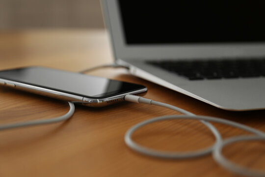 Modern smartphone charging from laptop on wooden table, closeup