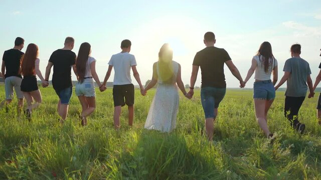 A large group of friends walks through the meadow holding hands in the evening at sunset.