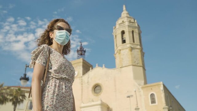 Beautiful curly brunette tourist with a surgical mask during Covid-19 with the Mediterranean village of Sitges on the bakground. Safe Travel in the new normal. Church of of Sant Bartomeu and Santa