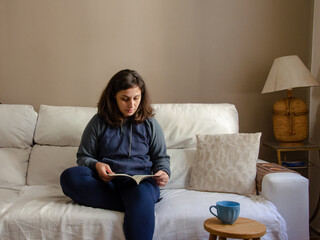 young woman sitting on a white sofa reading a book  using comfort clothes