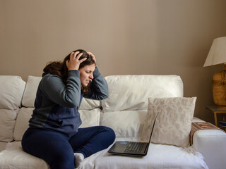 young woman sitting on a white sofa working on a laptop using comfort clothes looking worried with hands in head