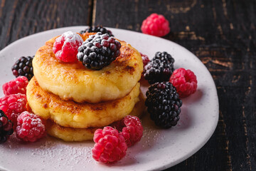 Cottage cheese pancakes and powdered sugar, curd fritters dessert with raspberry and blackberry berries in plate on dark black wooden background, angle view macro