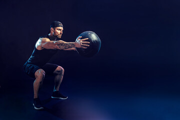 Aggressive bearded muscular sportsman is working out with a medicine ball isolated on dark studio background.