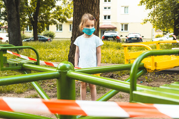 Little cute girl in face mask at closed children's playground. COVID-19 security and protection measures in the city. Concept of life and walking in new reality, lockdown, quarantine.