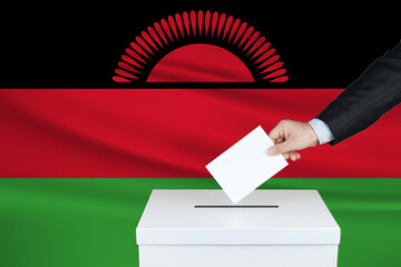 Election in Malawi. The hand of man putting his vote in the ballot box. Waved Malawi flag on background.                  
