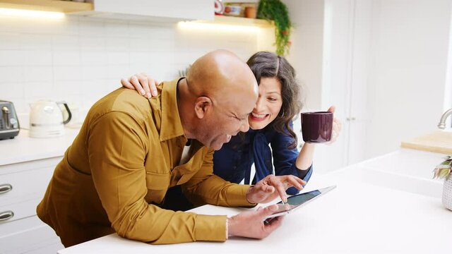 Mature couple at home in kitchen drinking coffee and looking at digital tablet together - shot in slow motion