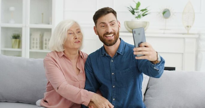 Caucasian young handsome man taking selfie photo with smartphone camera with senior beautiful gray-haired woman on sofa at home. Adult smiled son posing to phone with old mother in living room.