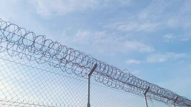 Low angle view of airport barbed wire security round metal fence on bright blue sunny sky day, handheld pan