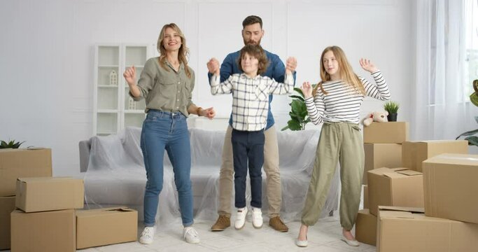 Happy cheerful Caucasian parents with small teen cute children standing in room of new house among carton boxes and smiling. Joyful kids, boy and girl, with mother and father moving in new home.