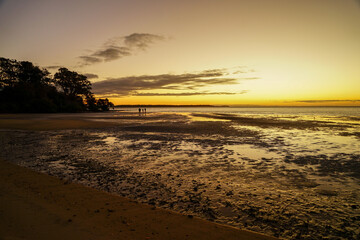 Golden glow of sunset on the beach at low tide, with silhouette of two people in the distance. Dunwich, North Stradbroke Island, Queensland, Australia.