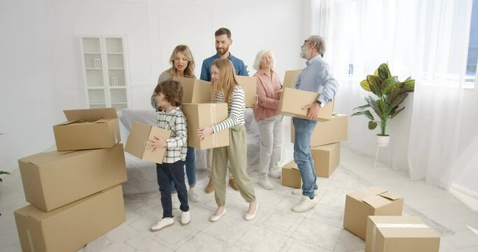 Caucasian happy family with kids and elder people coming in new home and carrying carton boxes. Mother, father, grandmother, grandfather, boy nd girl in house after repairment looking around Moving in