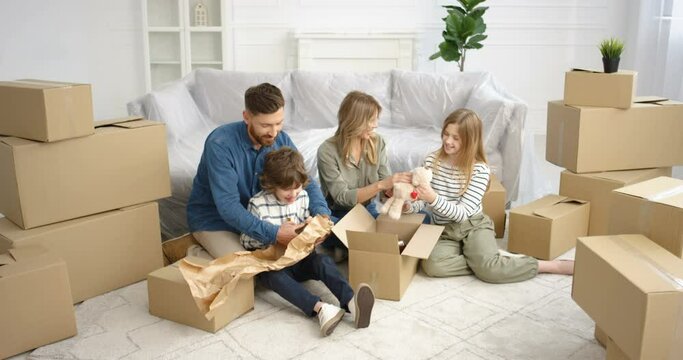 Cheerful Caucasian parents sitting on floor in living room with small cute kids and unpacking carton boxes. Family with children moving in new house after repairment Boy and girl with mother ad father