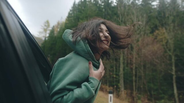 Close-up Of Asian Girl Traveler With Long Black Hair Traveling, Sitting In Front Seat Of Car, Crawling Out Of Window And Enjoying Wind. Woman Riding Car And Laughing, Her Face Cuting By Wind.