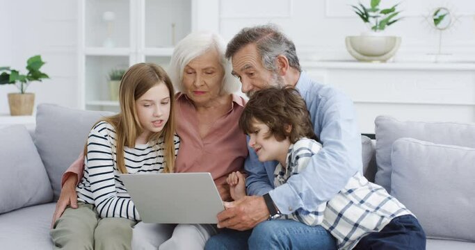 Caucasian senior grandparents with grandchildren sitting on couch at home and using laptop computer. Grandmother, grandfather, grandson anf granddaughter spending time together in internet. Indoors.