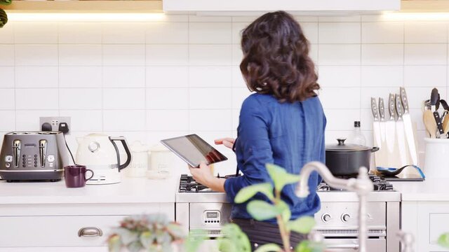 Mature woman at home in kitchen looking at digital tablet and boiling kettle for hot drink - shot in slow motion