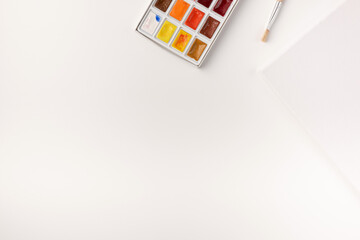 Watercolor paints, brushes, white canvas on white background. Copy space. Top view. Flat lay.