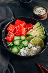 Hawaiian salmon poke bowl with cucumber, avocado and sesame seeds on black wooden table.