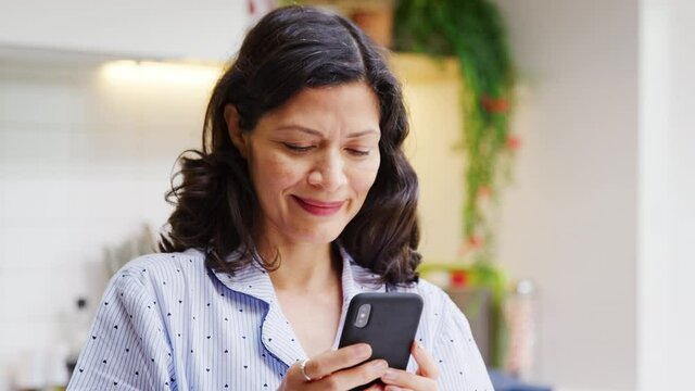Mature Hispanic woman in pyjamas at home in kitchen checking social media and text messaging on mobile phone - shot in slow motion 