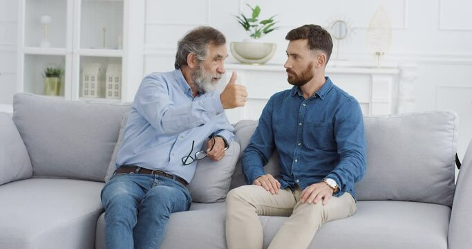 Caucasian males family generations. Happy cheerful senior father with gray hair telling something to adult son and explaining on couch in cozy living room. Old man teaching and preaching young guy.