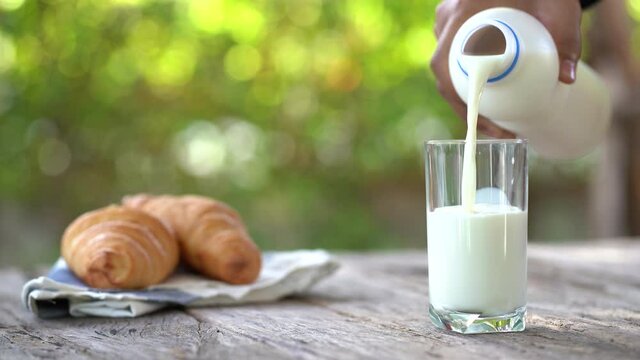 An empty glass and a bottle of milk stands on a wooden table. Girl's hand pours milk into a glass on a dark background.