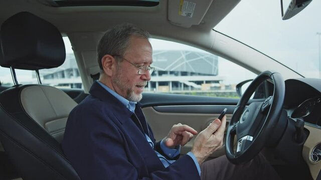 Bearded nice-looking senior businessman stops on the road, sitting in expensive modern car, reading messages from grandchildren and smiling with much joy. Successful entrepreneur. Retirement