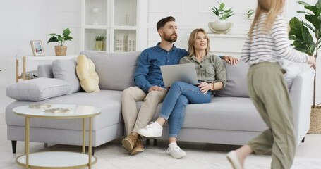 Two cheerful Caucasian kids running in circles and rounds in living room. Happy cute small sister and brother playing around sofa. Smiled parents sitting on couch with laptop computer Hugging children - Powered by Adobe