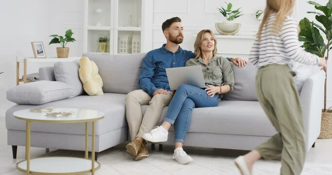 Two cheerful Caucasian kids running in circles and rounds in living room. Happy cute small sister and brother playing around sofa. Smiled parents sitting on couch with laptop computer Hugging children