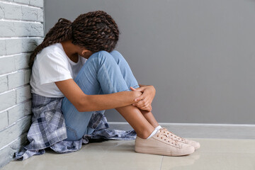 Sad African-American girl sitting near grey wall. Stop racism